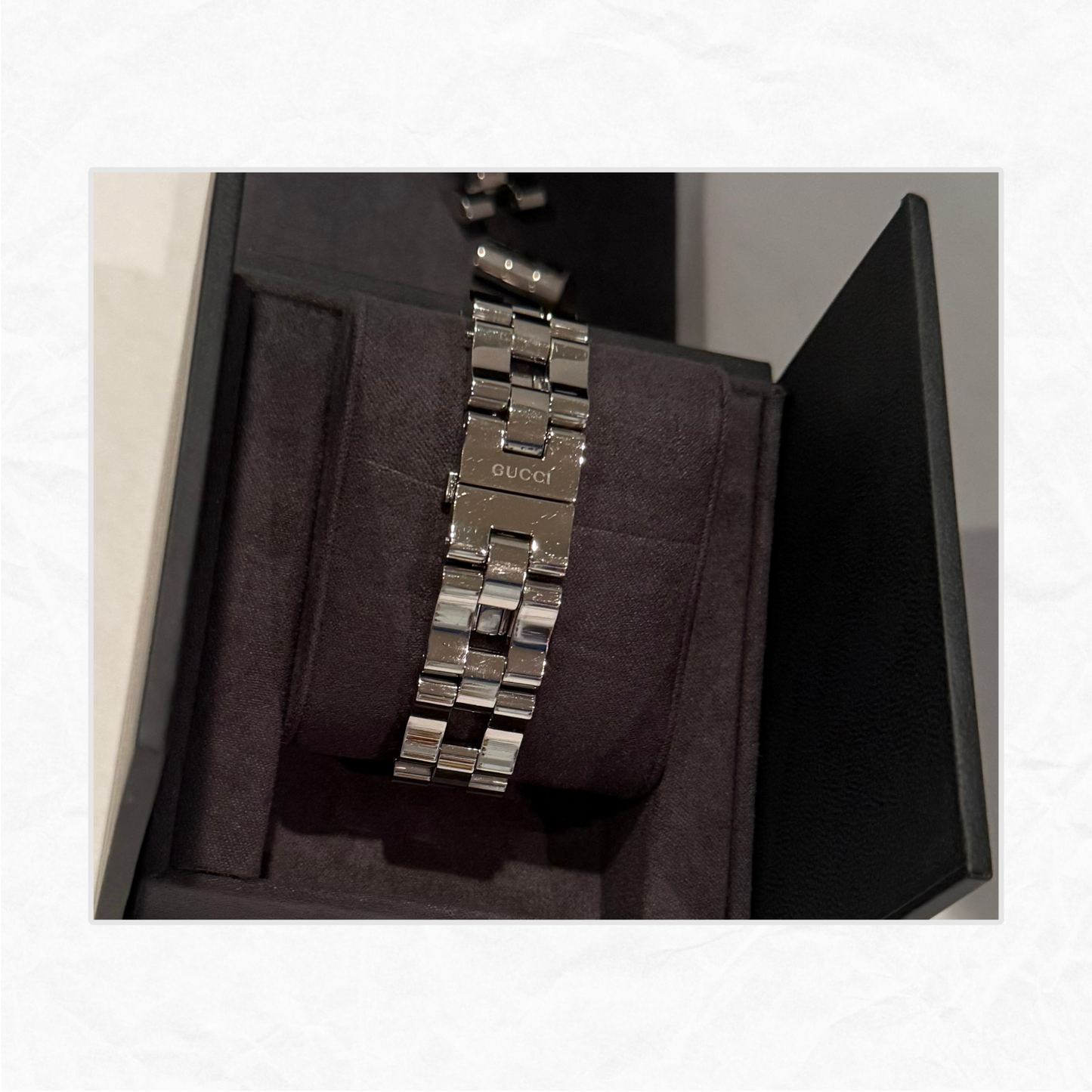 A silver stainless steel Gucci link watch band displayed inside a black watch box with a dark velvet cushion. The brand name 'GUCCI' is engraved on the clasp.